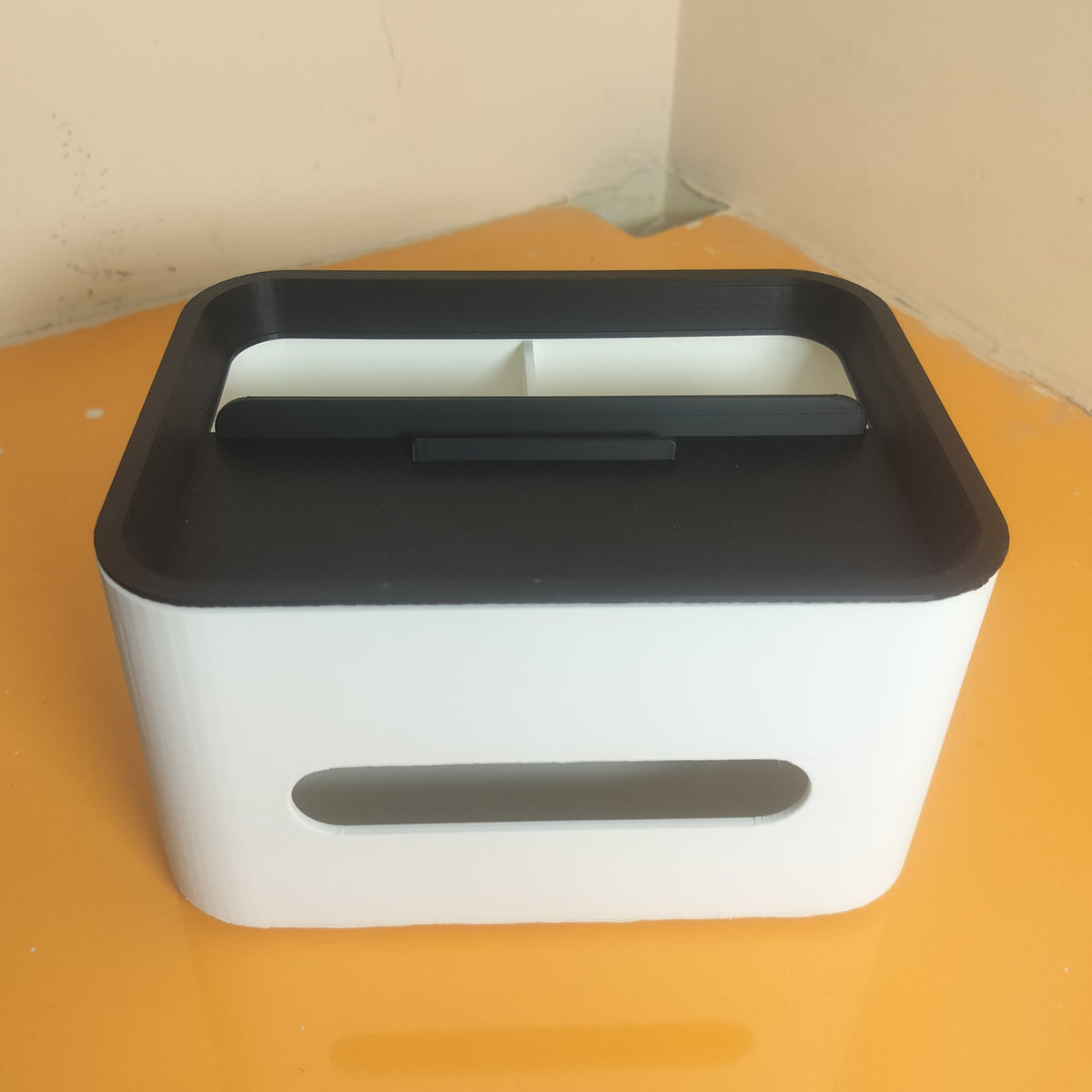A white and black plastic tissue box cover or small organizer sitting on a yellow-orange surface against a beige wall corner.