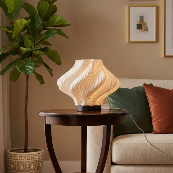 A modern sculptural table lamp with a fluted, pleated white shade and a black circular base, sitting on a glossy orange surface against a beige wall.
