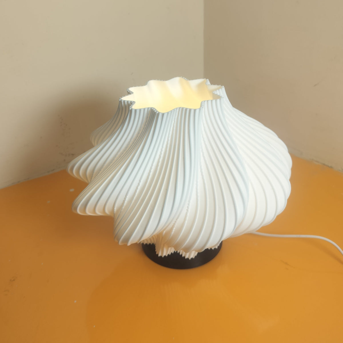 A modern sculptural table lamp with a pleated, flower-like white shade, glowing from within, set on a yellow surface against a beige wall.