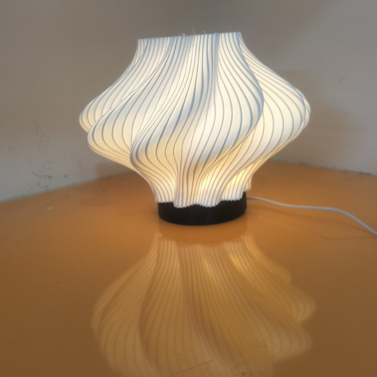 A modern pleated table lamp with a white and blue striped shade glowing with warm light, sitting on a reflective orange surface against a plain gray wall.