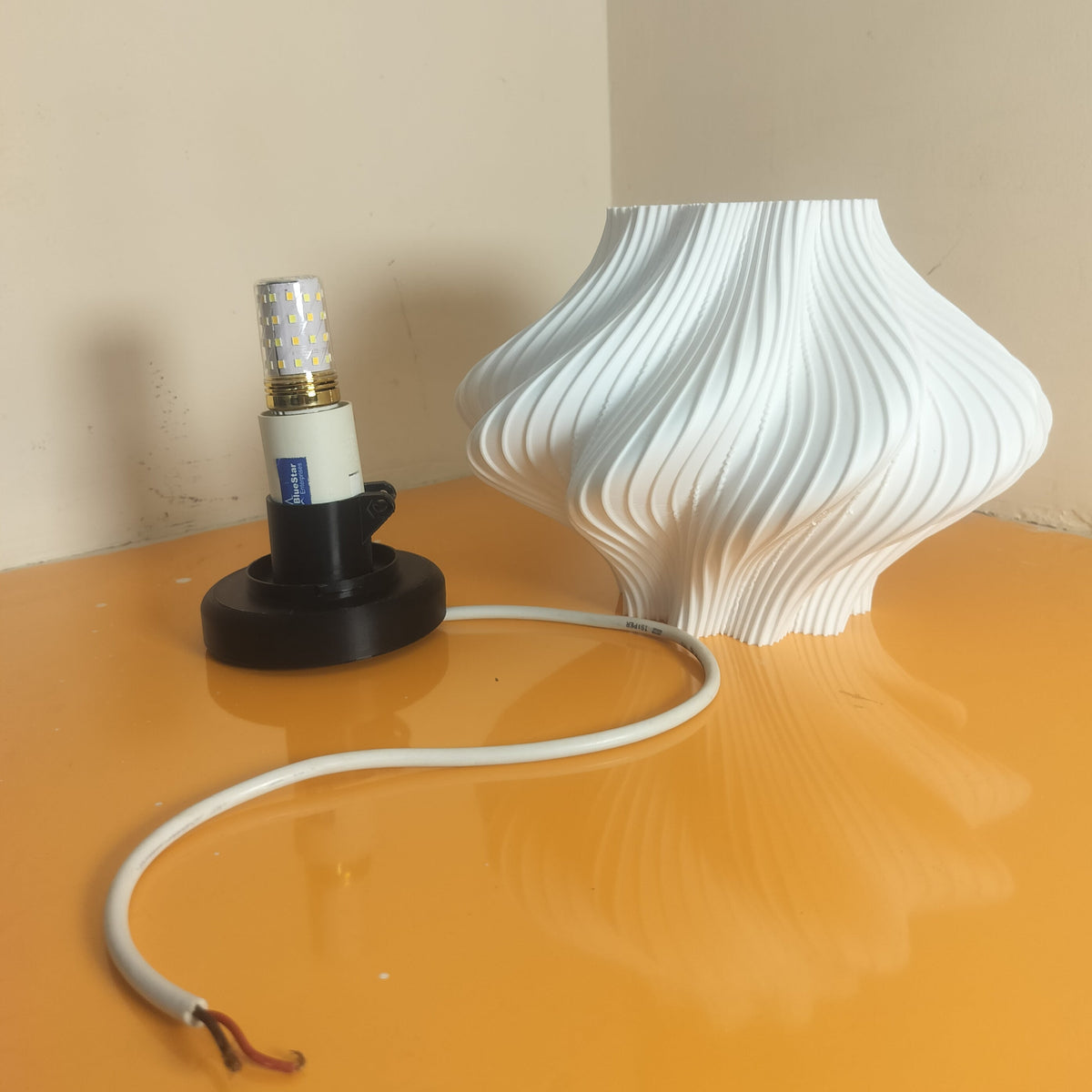 A white ribbed, bulb-shaped lamp shade or decorative vase sits on a glossy orange surface next to a black lamp base with a white cord, against a beige wall corner.