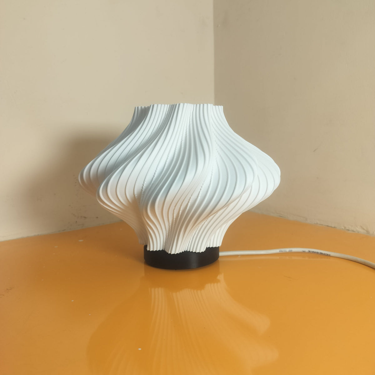 A modern white ribbed lamp with a black circular base sits on a glossy orange table against a beige wall.