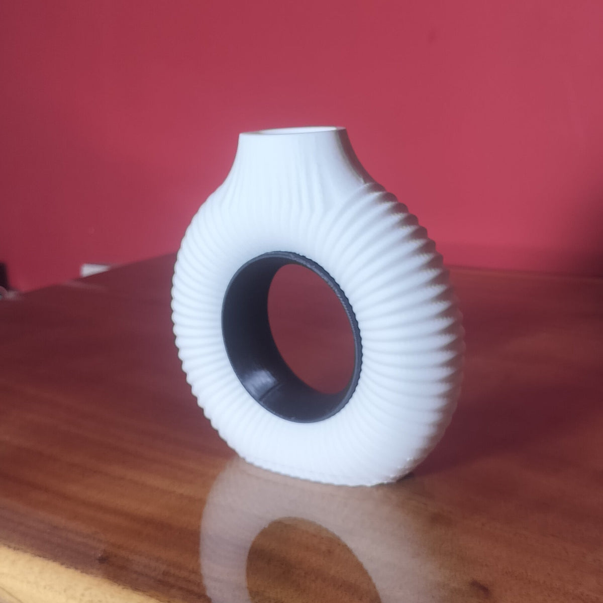 A white ribbed ceramic vase with a circular cutout, displayed on a polished wooden table against a solid red wall.