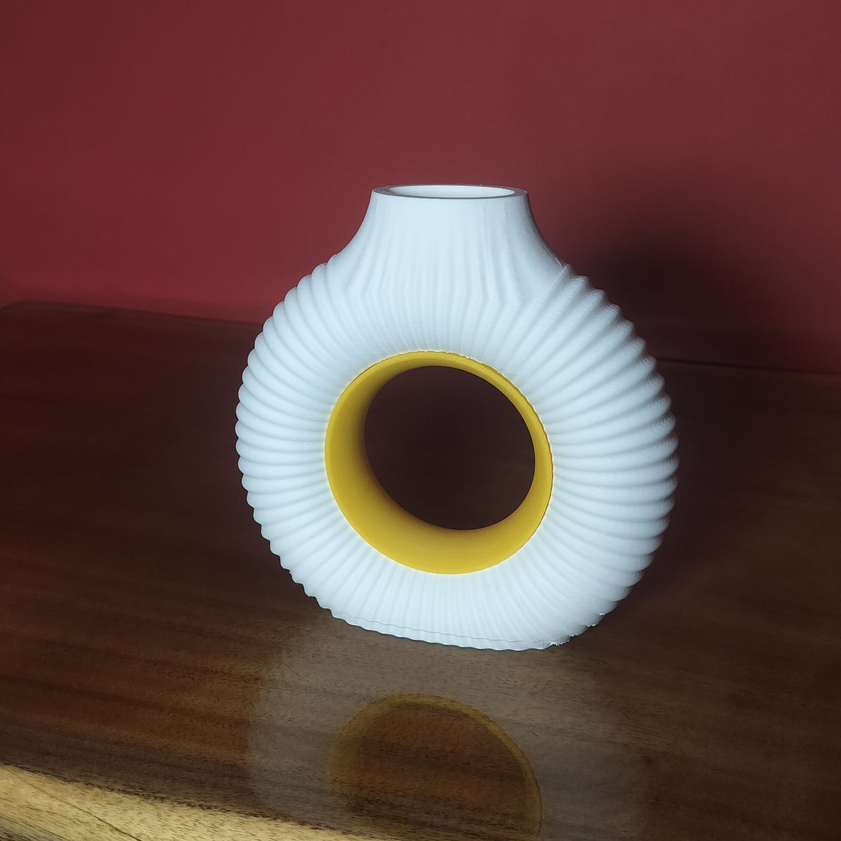 A white ribbed ceramic vase with a yellow inner ring, displayed on a polished wooden table against a dark red wall.