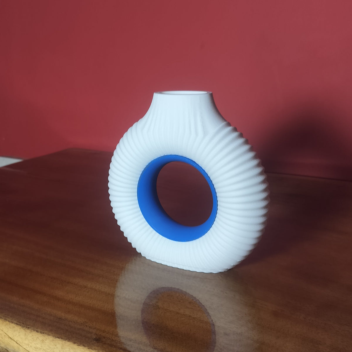 A white ribbed ceramic vase with a blue inner ring, displayed on a polished wooden table against a red wall.