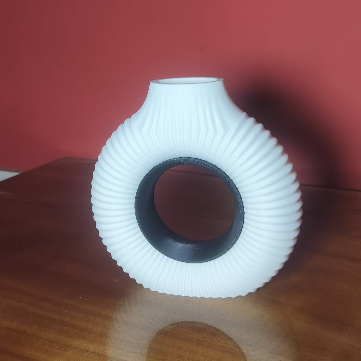 A white ribbed ceramic vase with a circular center hole, displayed on a wooden table against a red wall.