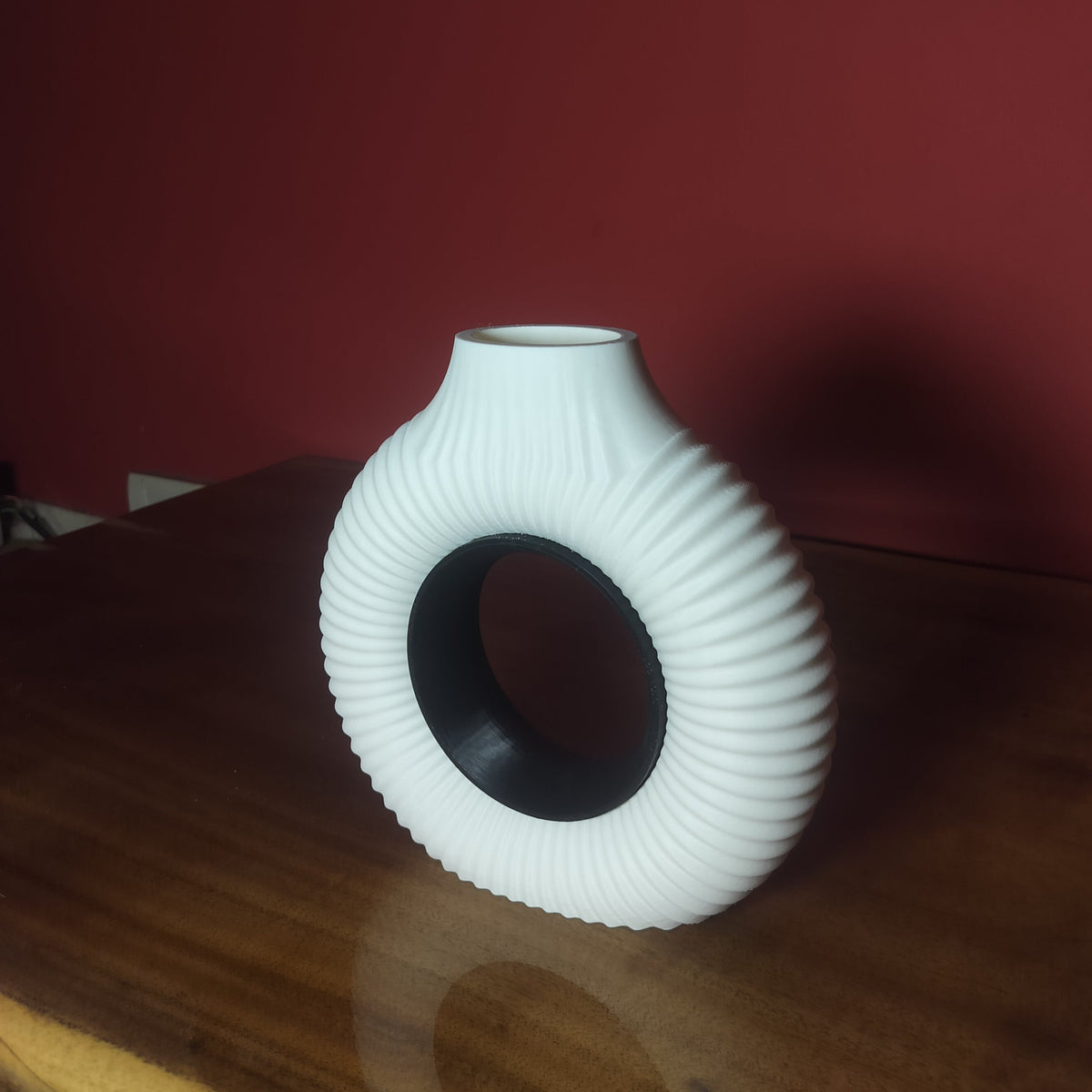 A white ribbed  vase with a hollow center, standing on a wooden table against a dark red wall.