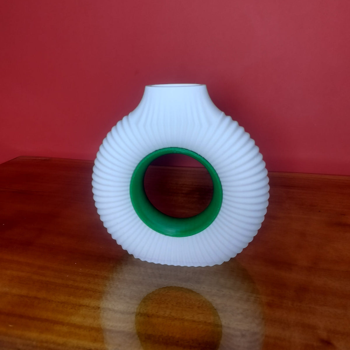 A white vase with ring-shaped insert with a green inner ring, standing on a wooden surface against a solid red background.
