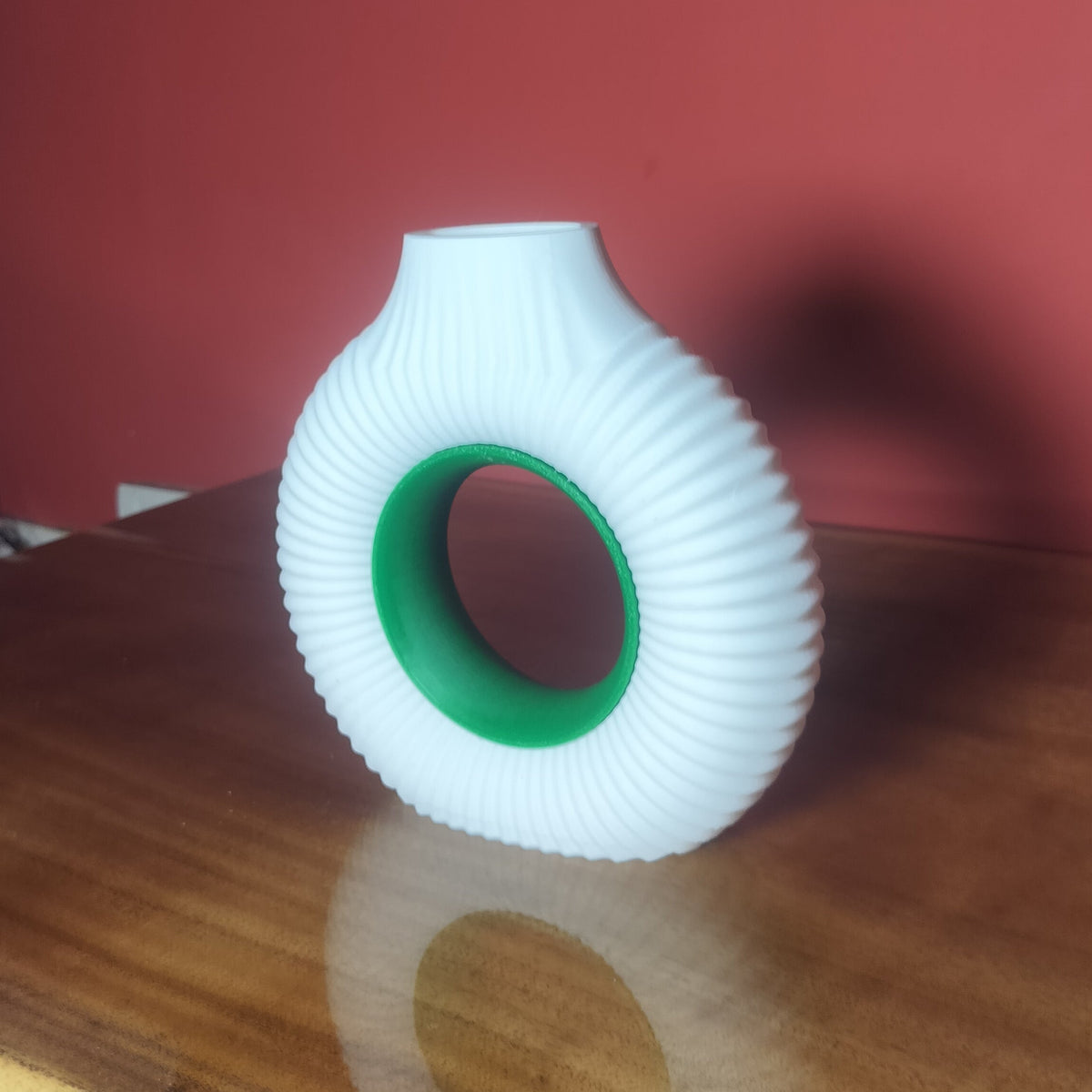 A white ribbed vase with a green inner ring, standing on a wooden table against a red wall.