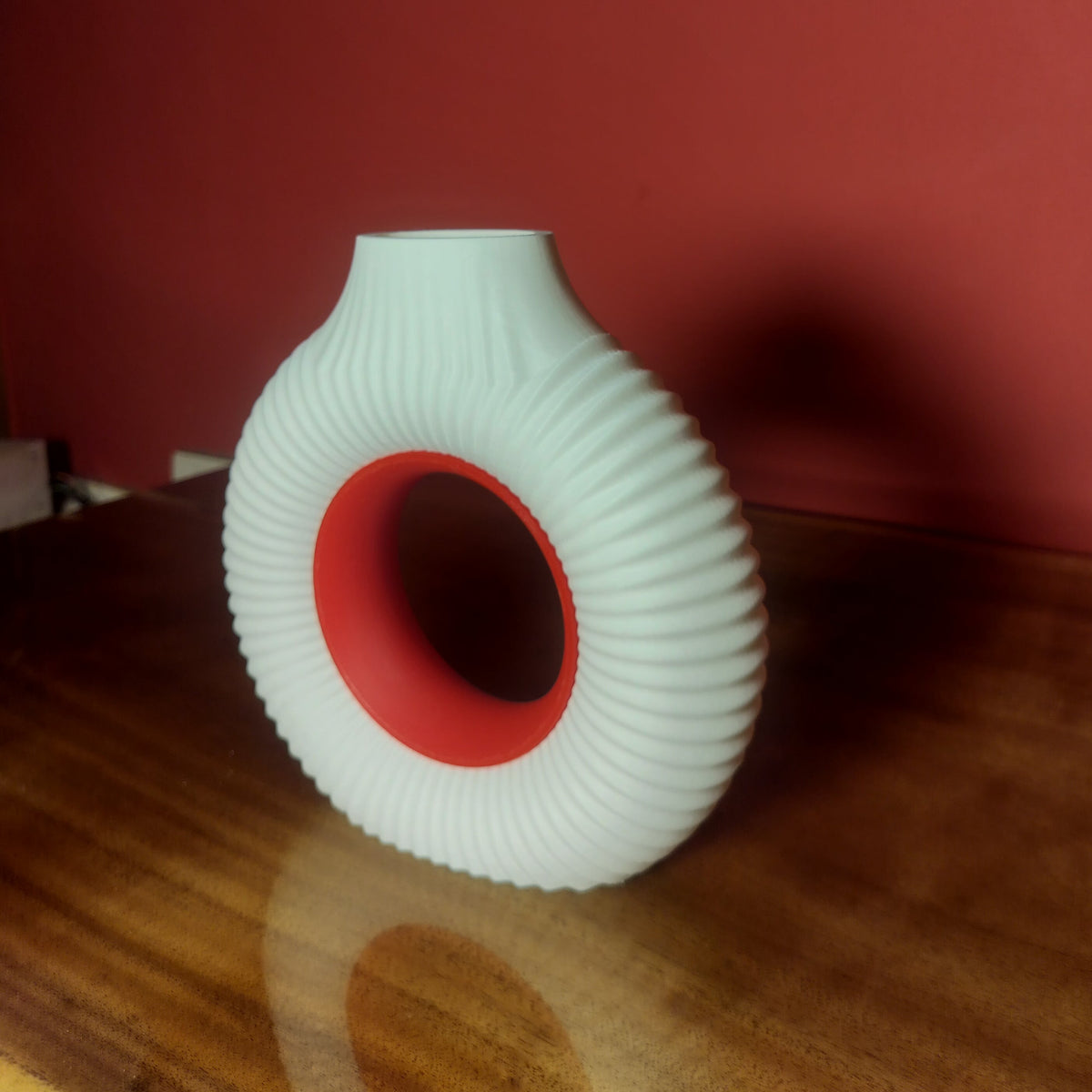 A ribbed white donut vase with a red interior, standing on a wooden table against a dark red wall.