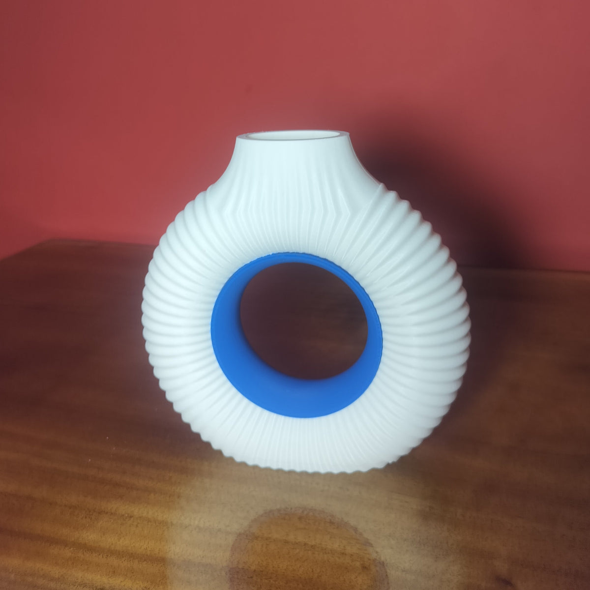 A white ribbed ceramic vase with a blue inner ring, displayed on a wooden surface against a solid red background.
