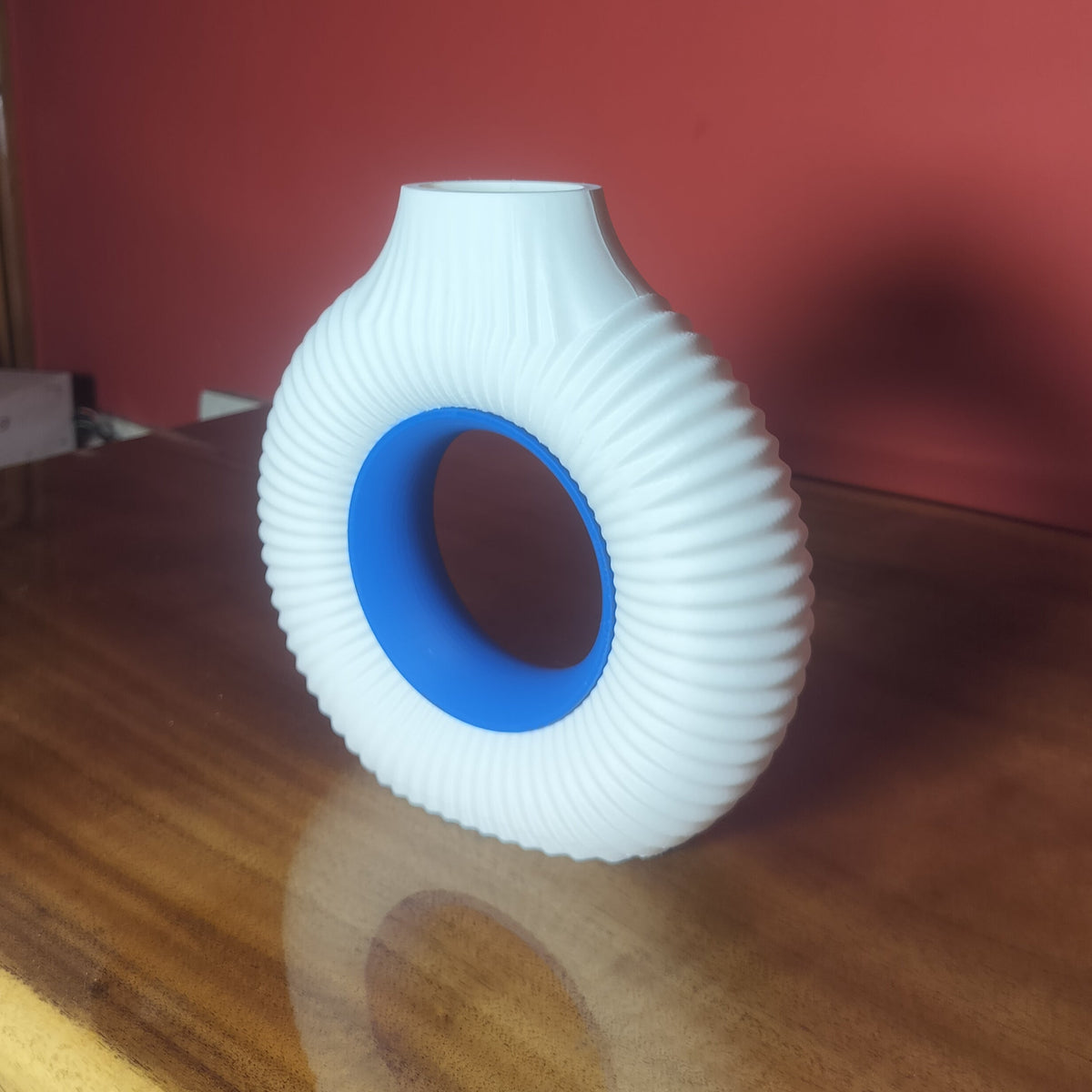 A white ribbed ceramic vase with a blue inner ring, standing on a wooden table against a red wall.
