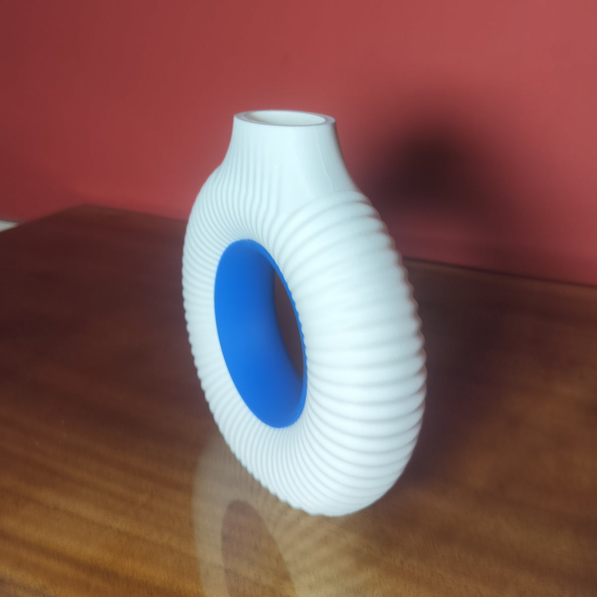 A white ribbed  ring-shaped vase with a blue center, standing on a wooden table against a red wall.