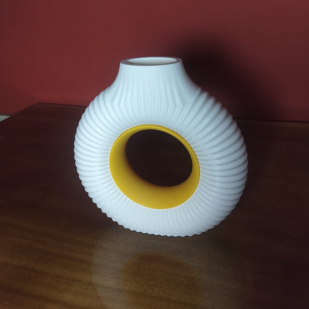 A white ribbed ceramic vase with a yellow inner ring, standing on a dark wooden table against a dark red wall.