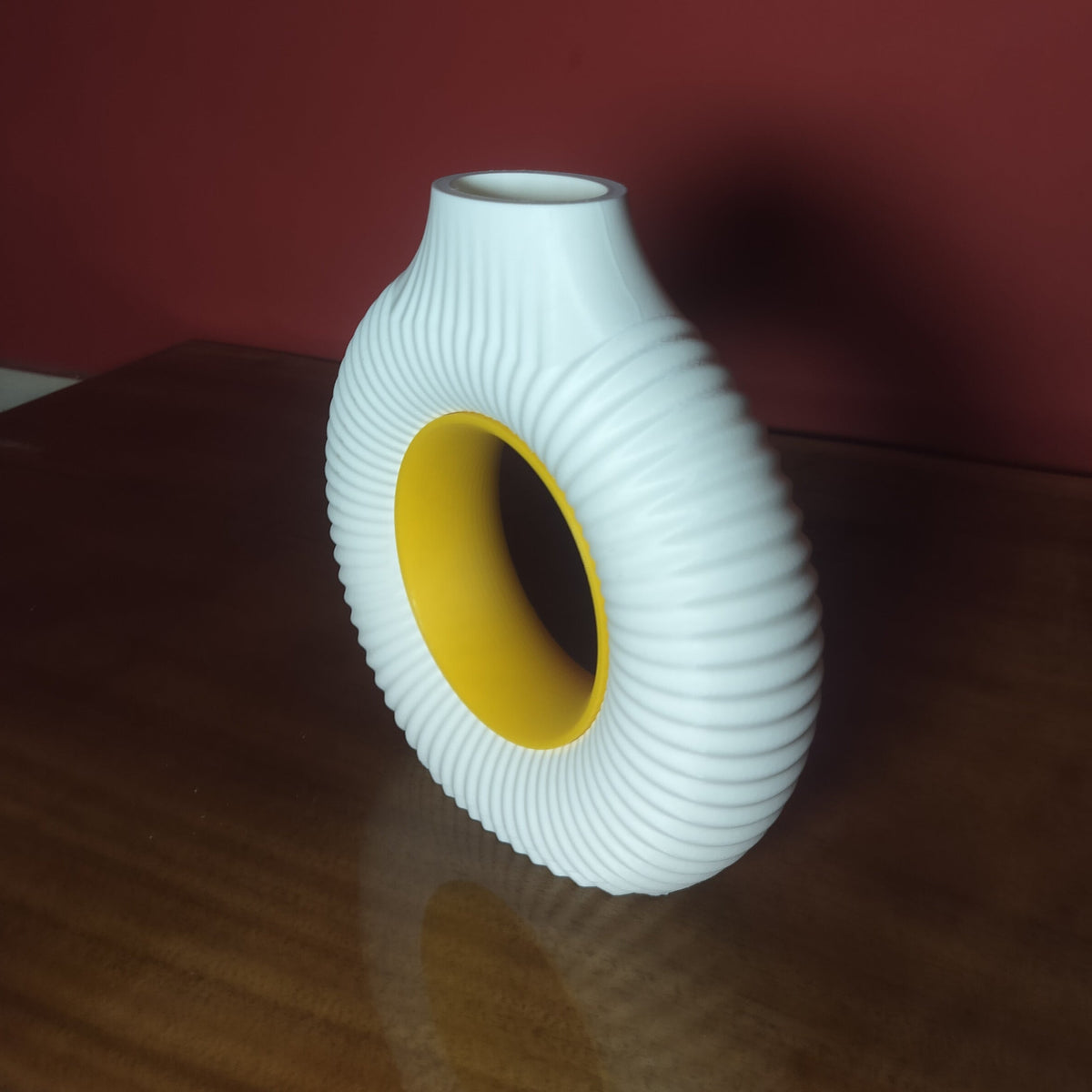 A white ribbed ceramic vase with a hollow center, showing a yellow interior, standing on a dark wooden table against a dark red wall.