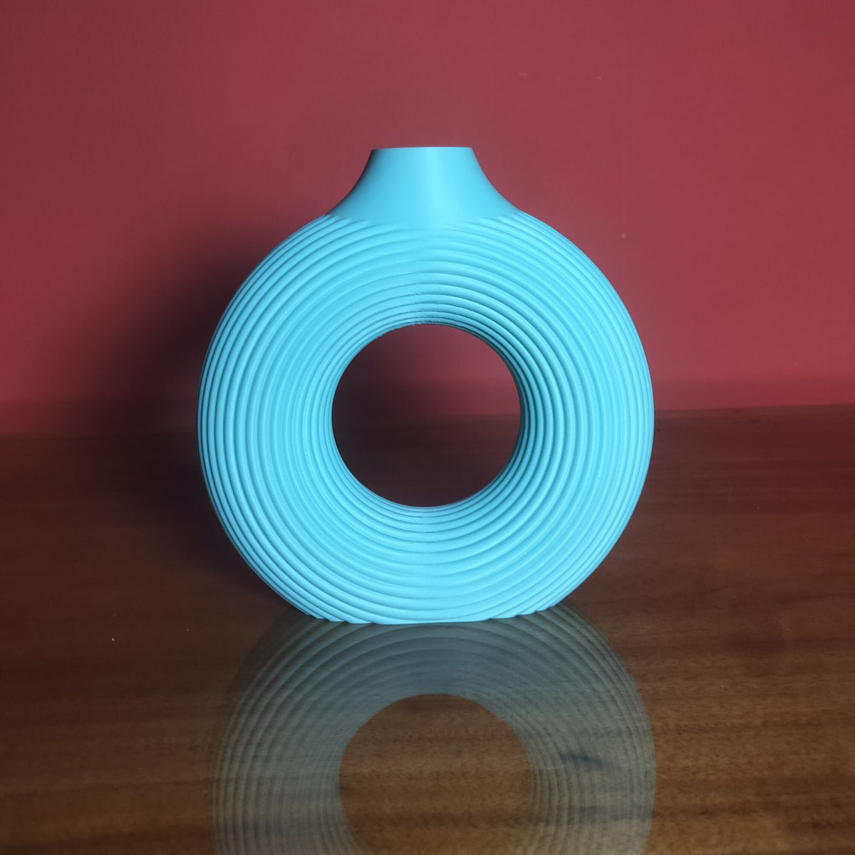 A light blue silicone ring-shaped personal massager with a flared base, standing upright on a reflective wooden surface against a solid dark red background.