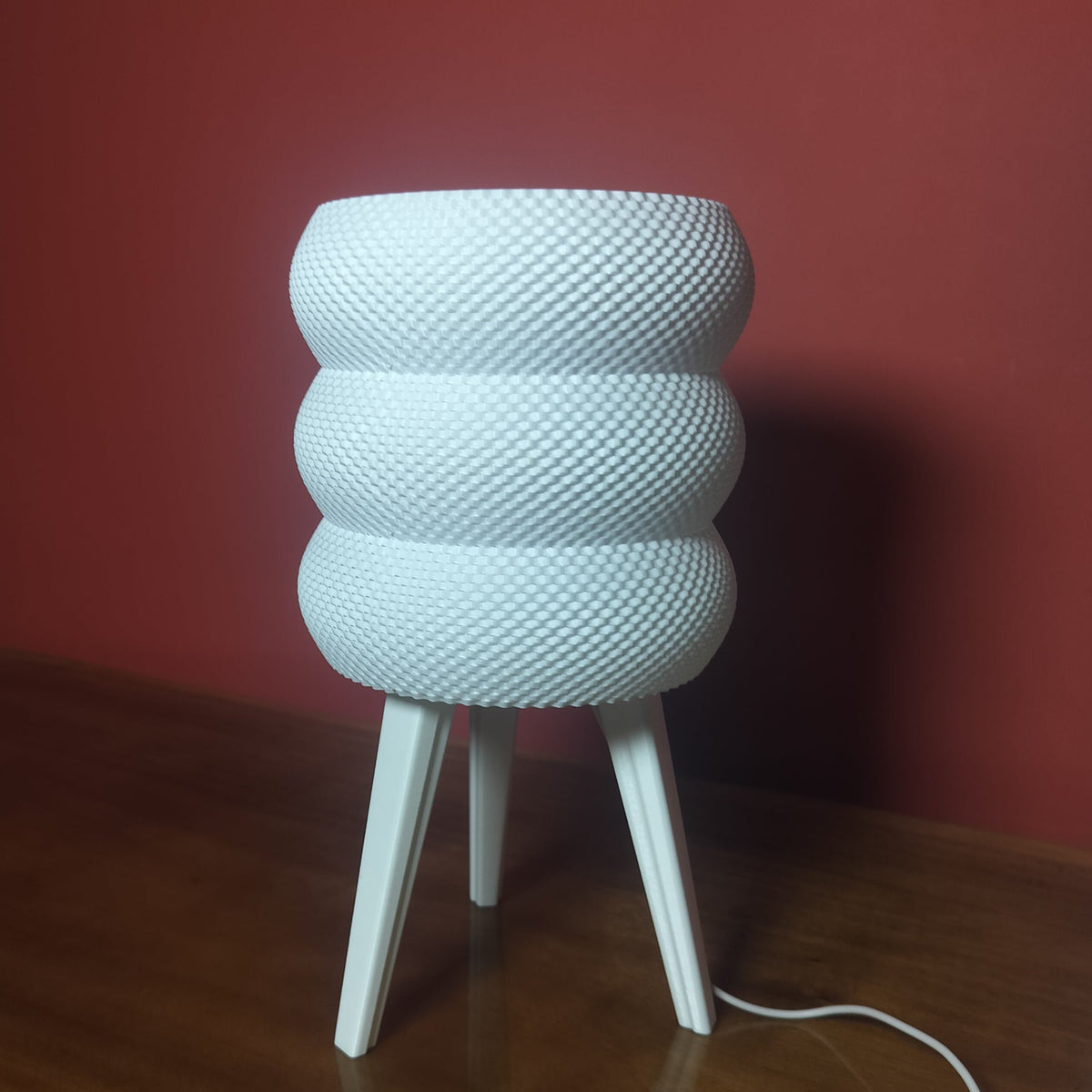 A white textured table lamp with a ribbed, rounded cylindrical body and three tapered legs, standing on a wooden table against a dark red wall.