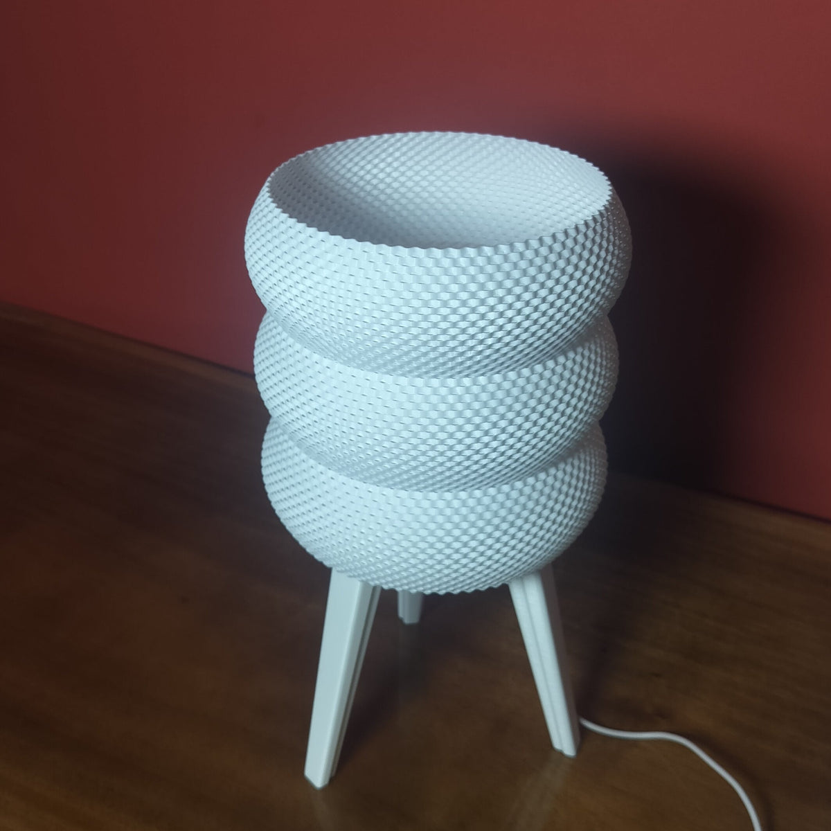 A white textured three-tiered bowl lamp with three legs, sitting on a wooden table against a dark red wall.