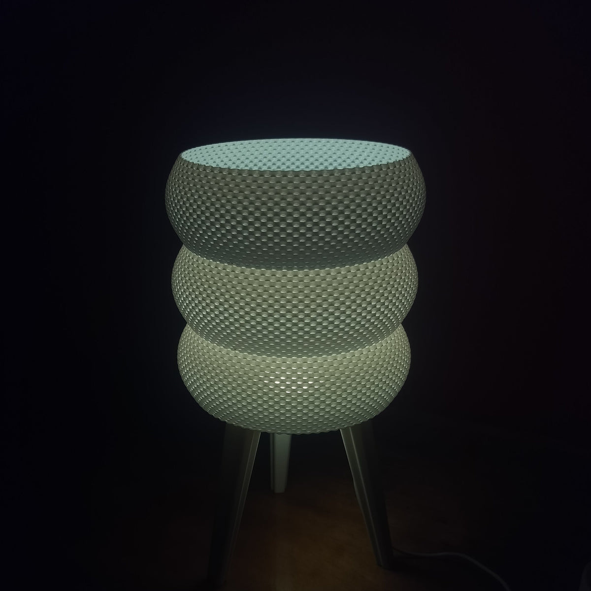A modern table lamp with three stacked, textured spherical shades on slim metal legs, glowing with a cool white light against a dark background.