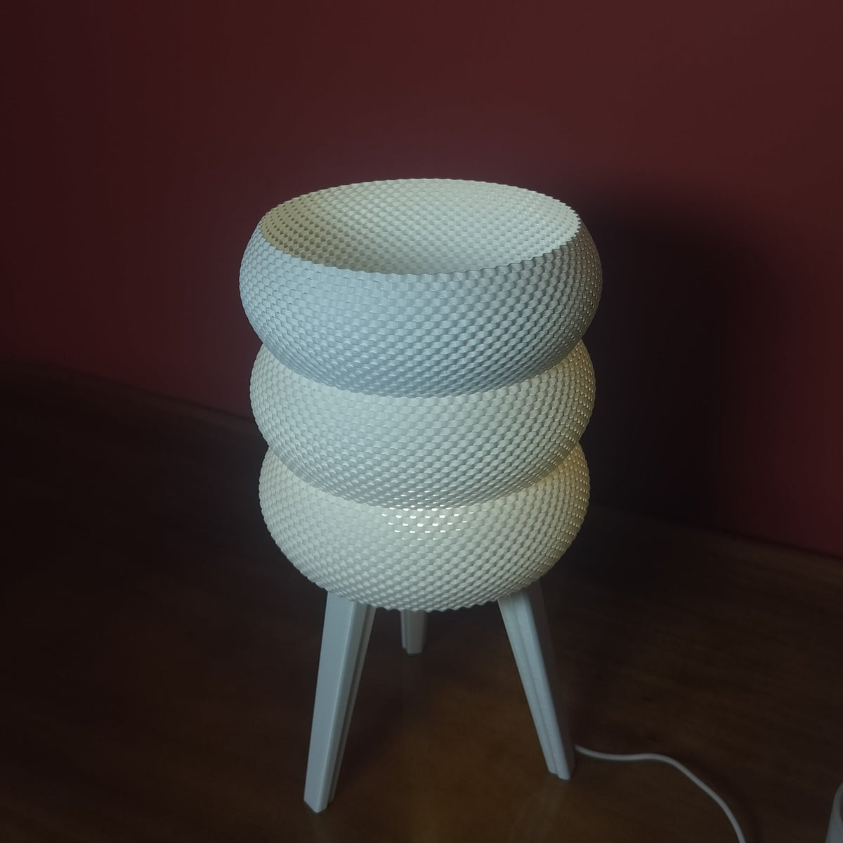 A white textured three-tiered bowl lamp with three legs, sitting on a wooden table against a dark red wall.