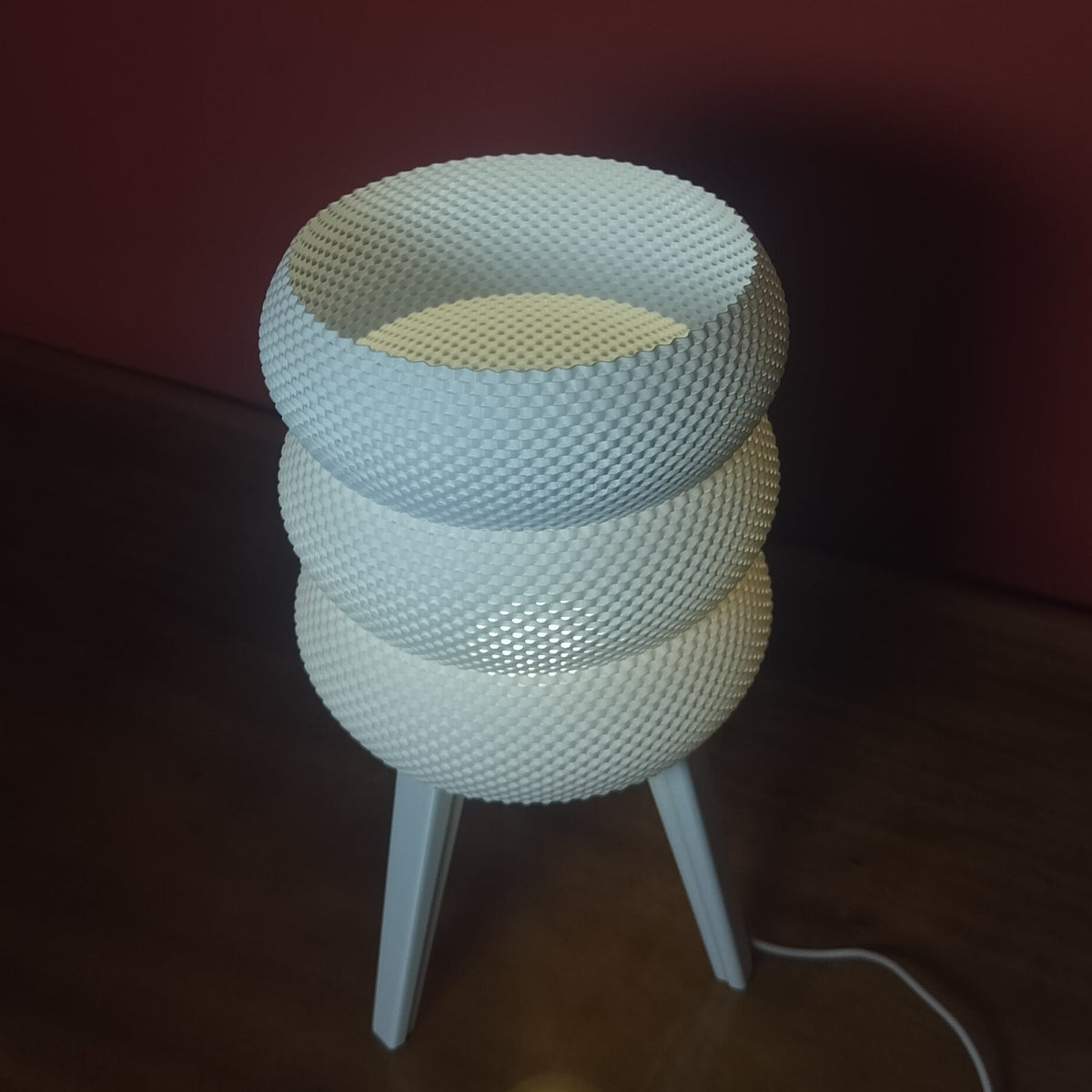 A modern white textured table lamp with a stacked, coiled design on three slim legs, sitting on a dark wooden table against a dark red wall.