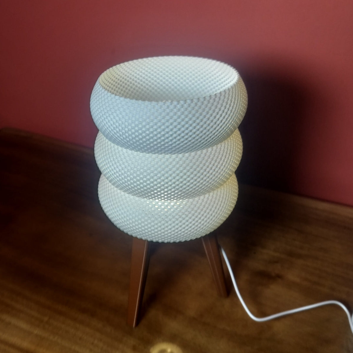 A modern table lamp with a textured, ribbed white circular shade stacked in three tiers on a wooden tripod base, set against a dark red wall and wooden tabletop.