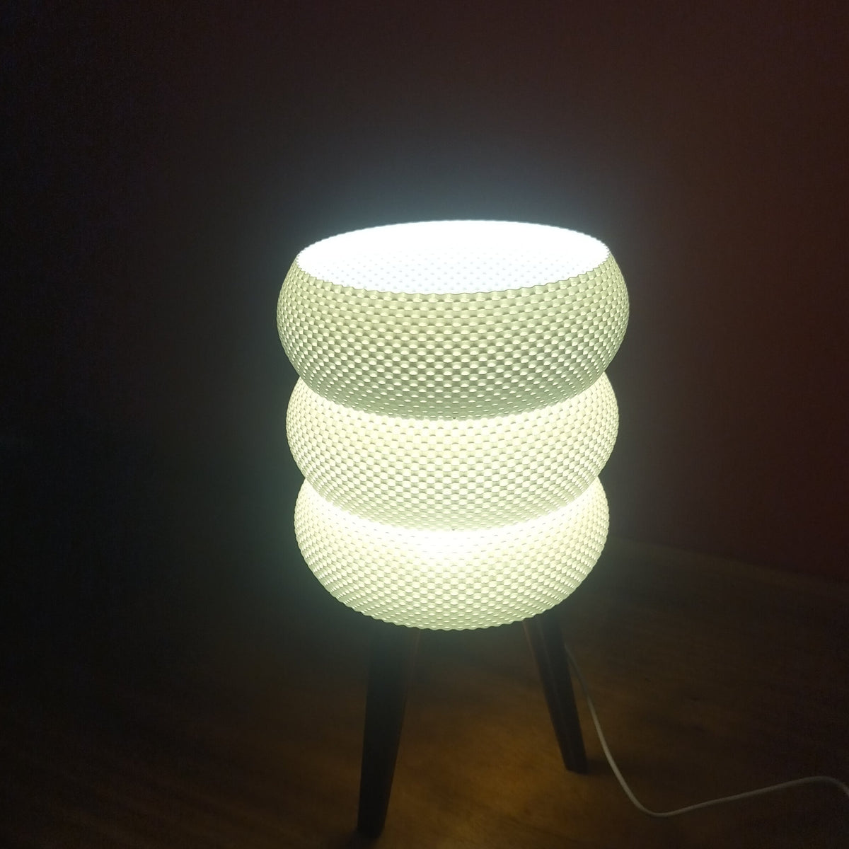 A glowing table lamp with three stacked textured white shades on thin dark legs, placed on a wooden surface against a dark background.