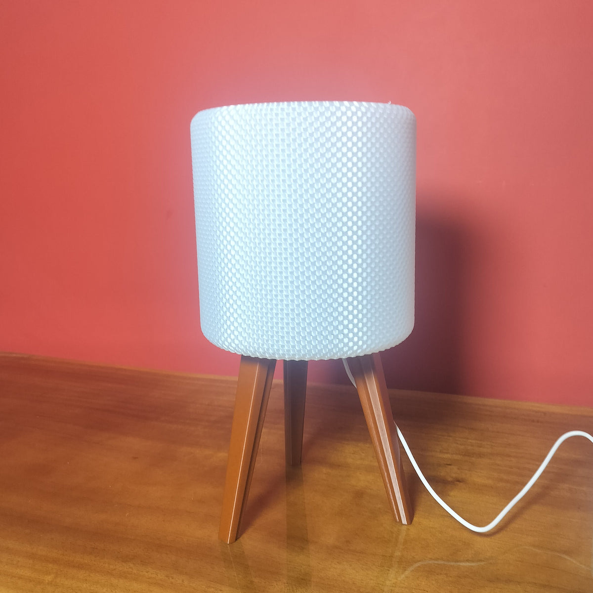 A modern table lamp with a white textured cylindrical shade and three angled wooden legs, sitting on a wooden tabletop against a solid red wall.