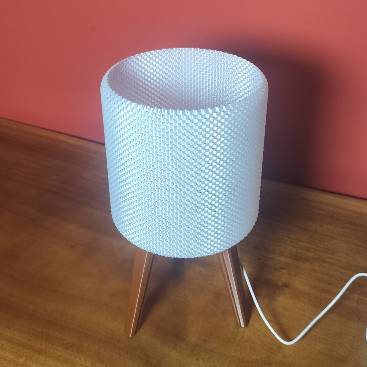 A modern table lamp with a white textured cylindrical shade and three wooden legs, sitting on a wooden table against a red wall.