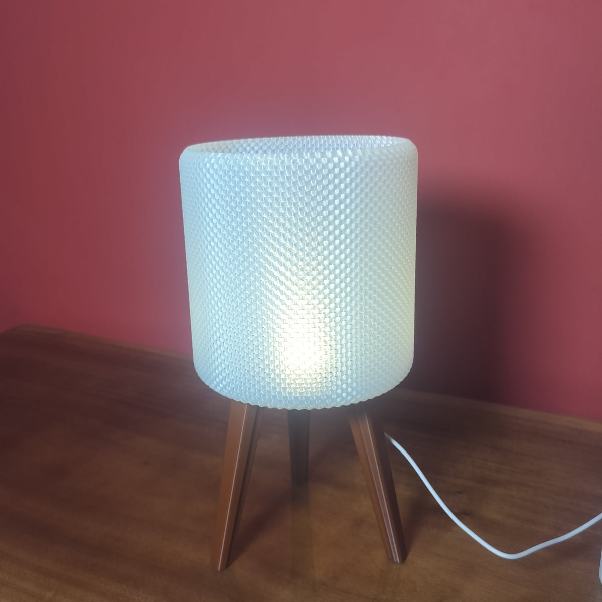 A modern table lamp with a textured white cylindrical shade and three wooden legs, illuminated and placed on a wooden table against a red wall.