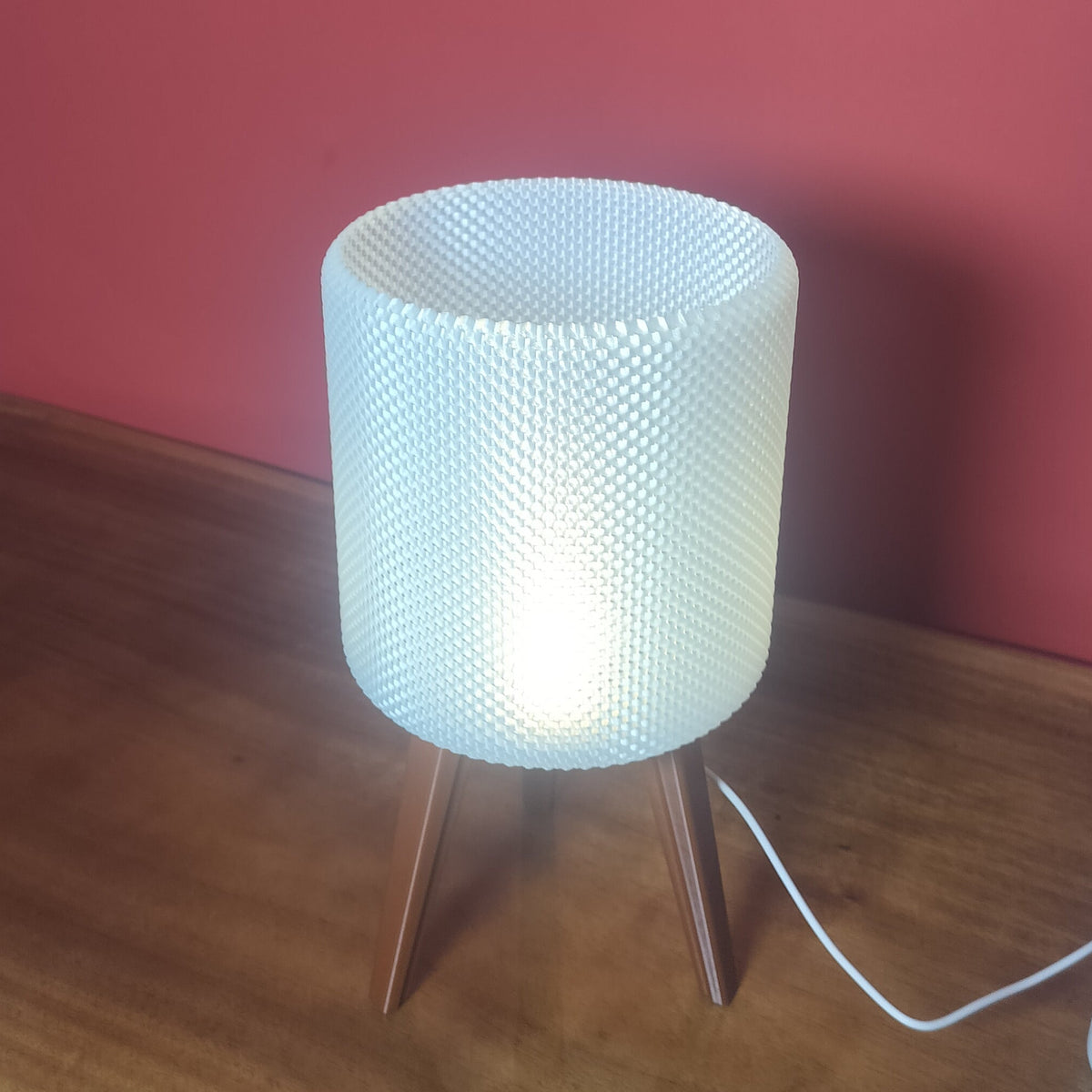 A modern table lamp with a glowing white textured cylindrical shade and three wooden legs, sitting on a wooden table against a red wall.
