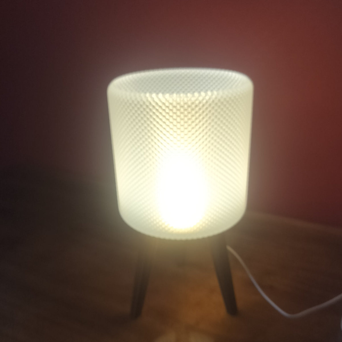 A glowing table lamp with a textured cylindrical shade and three short legs, placed on a wooden surface next to a small candle holder, against a dark red wall.