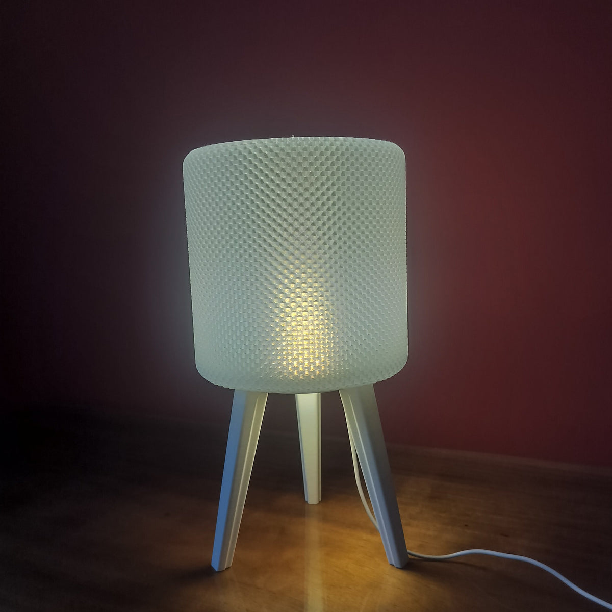 A modern table lamp with a textured cylindrical shade and three angled legs, glowing with warm light on a wooden surface against a dark reddish-brown background.