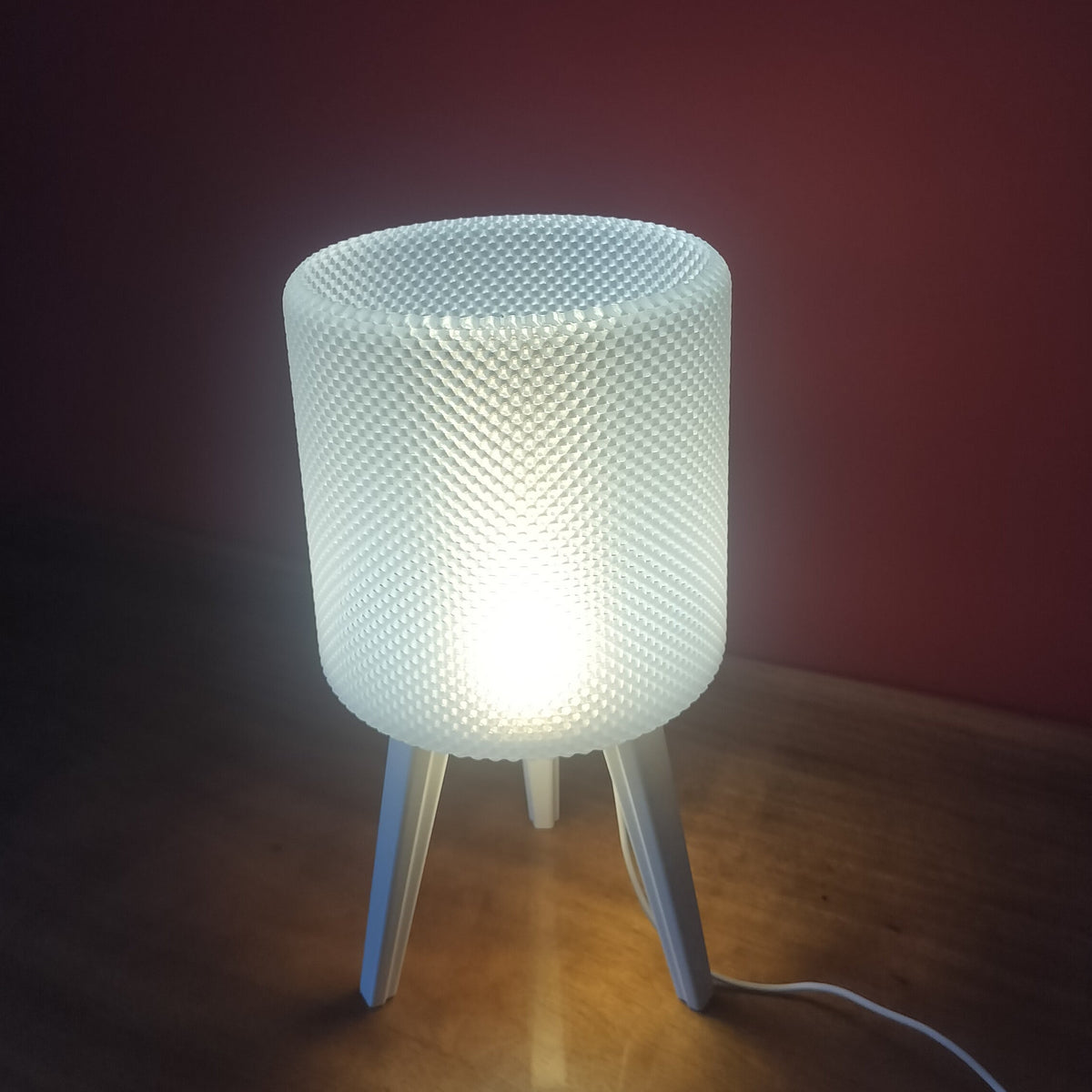 A modern table lamp with a cylindrical, textured white shade supported by three slim legs, glowing with a warm light on a wooden surface against a dark red wall.