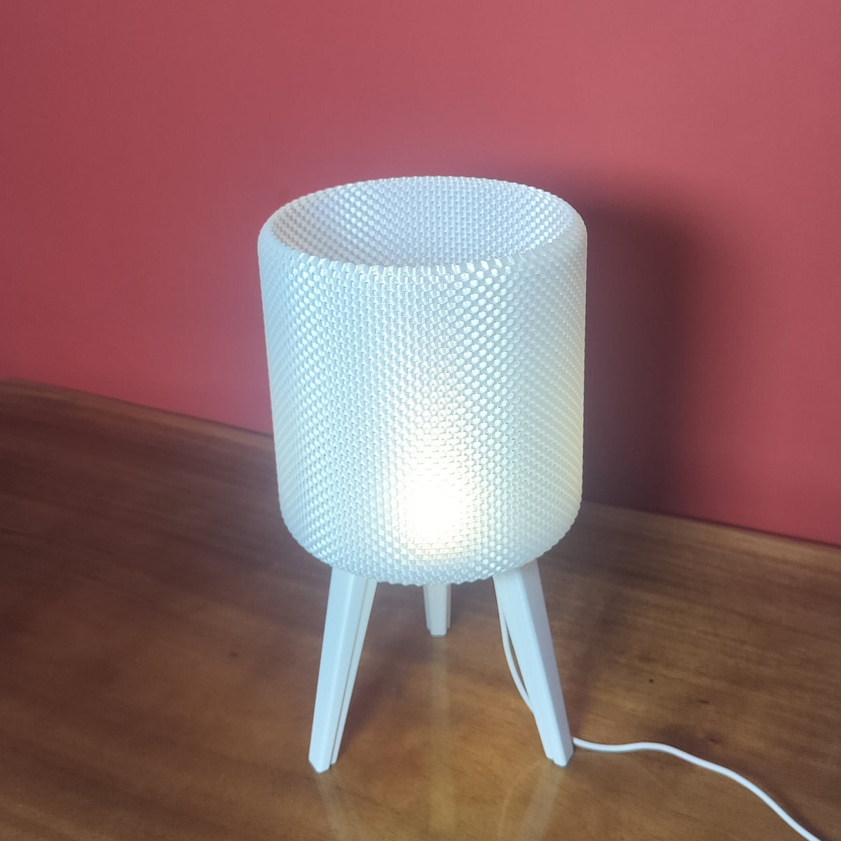 A modern white table lamp with a textured cylindrical shade and three tapered legs, illuminated on a wooden table against a red wall.