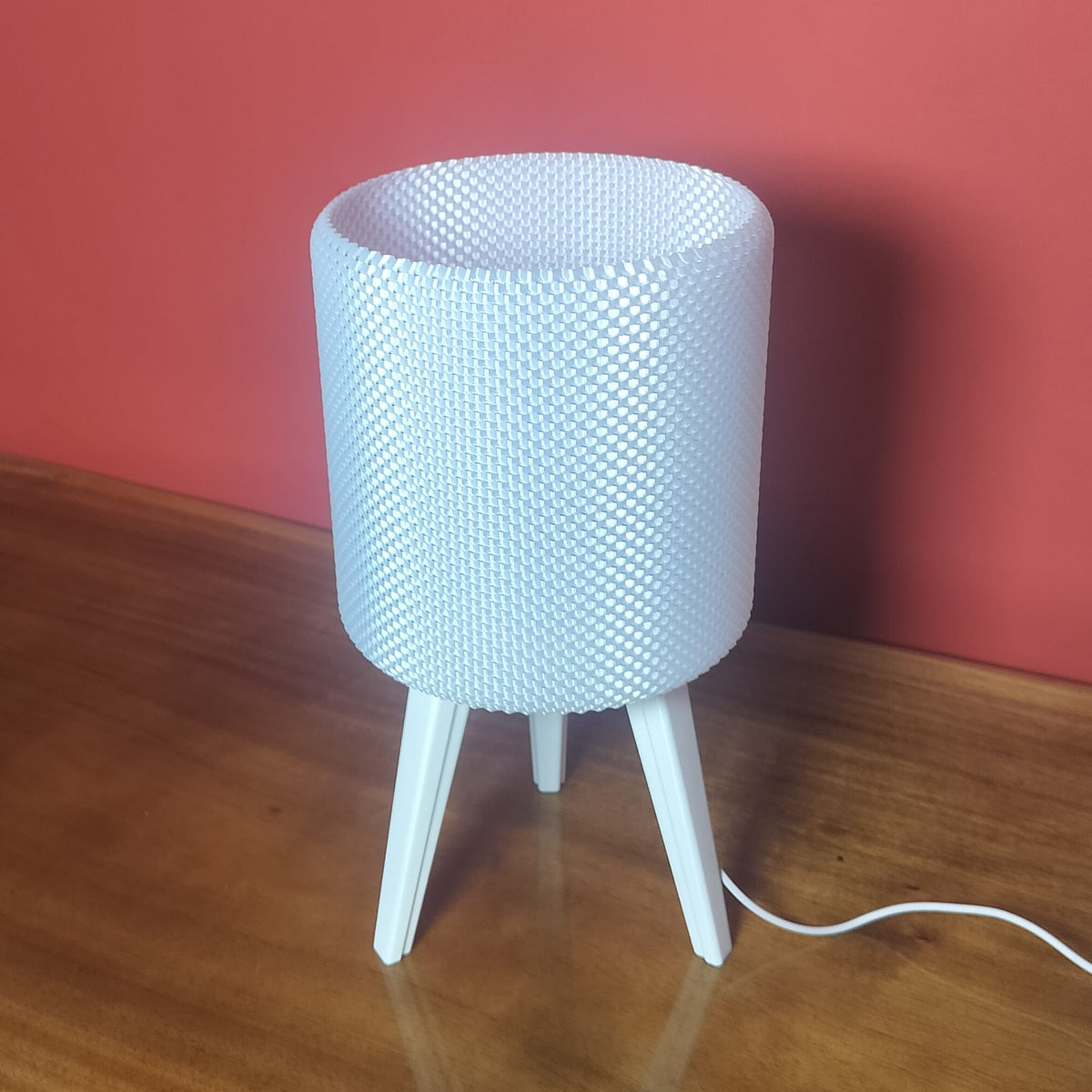 A white textured cylindrical table lamp with three legs sits on a wooden table against a red wall.