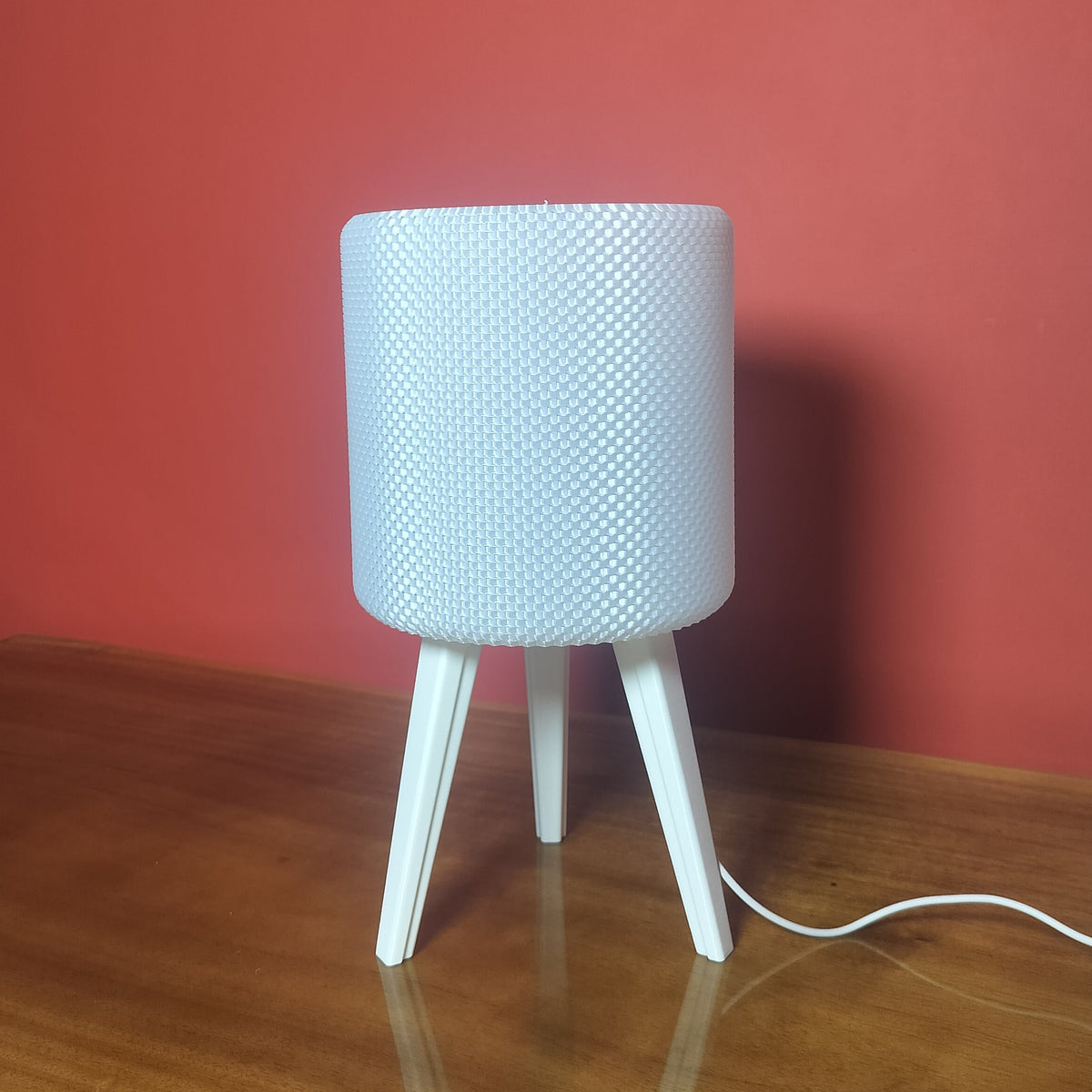 A modern white table lamp with a textured cylindrical shade and three tapered legs, sitting on a wooden table against a solid red wall.